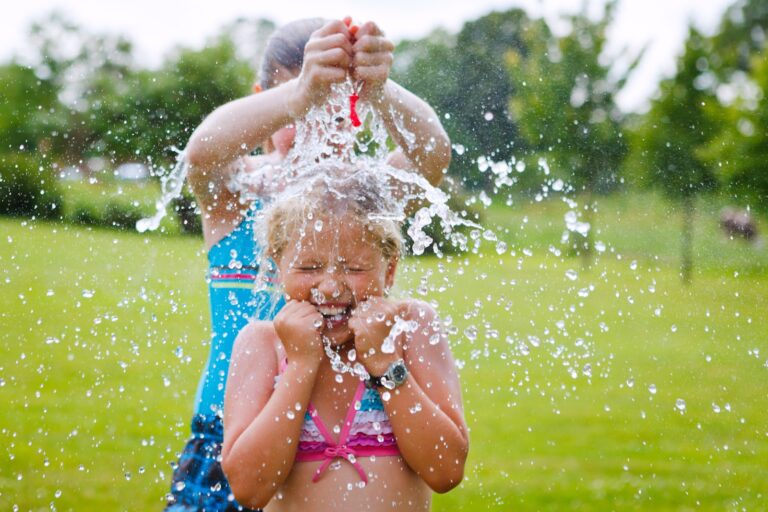 Girl splashes water onto a smaller girl during the summer in a large field of grass.
